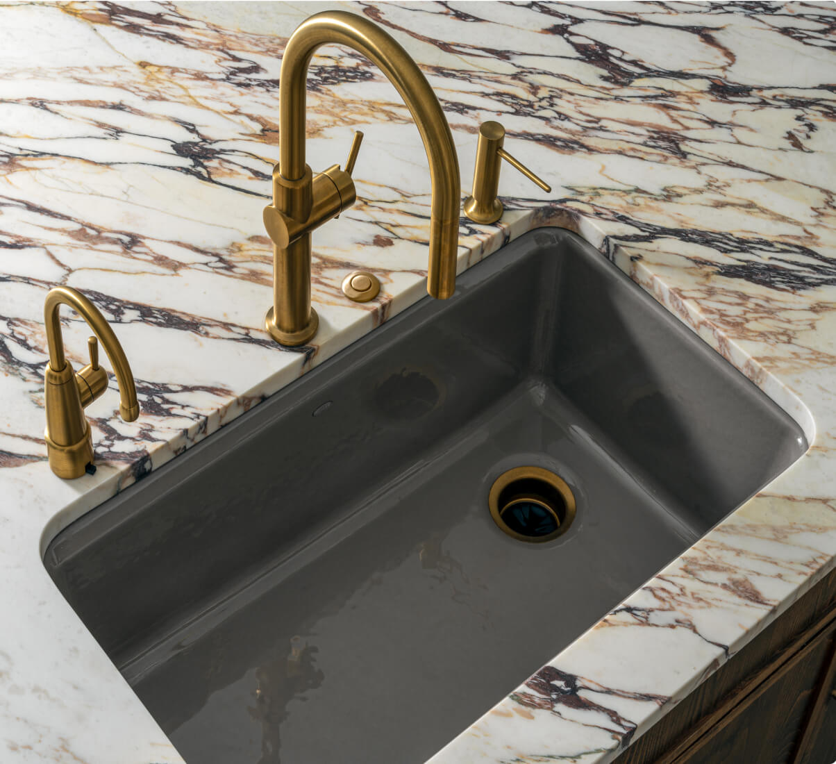 A close up of a grey ceramic sink in a marble island with black and gold veining. The faucet is also in gold.