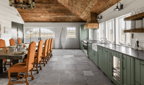 Open kitchen with green cabinetry, stone flooring, custom wood vent hood and a spacious dining table for entertaining.